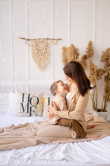 mom gently hugs a little baby boy at home on a bed in natural brown tones, a happy family with a small child has fun together