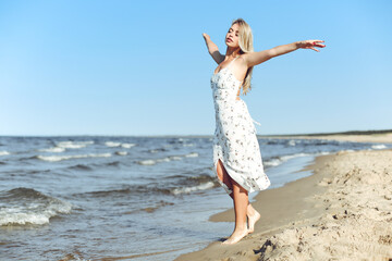 Fototapeta na wymiar Happy blonde beautiful woman on the ocean beach standing in a white summer dress, open arms.