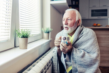 Elderly man in living room using heater at home in winter. Senior man warming his hands. Heating...