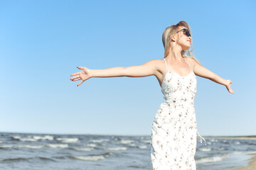 Fototapeta na wymiar Happy blonde beautiful woman on the ocean beach standing in a white summer dress and sun glasses, open arms.