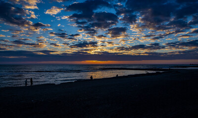 Obraz premium Some people take a walk on the beach of Ostia, Rome, in the sunset, in winter. The sun covered by clouds and the reflection of warm light on the blue sea water.