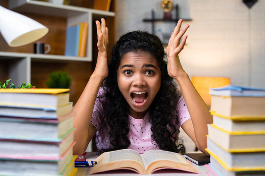 Shouting or screaming young girl in front of stack of books on study table at home - concept of exam stess, depression and education