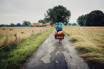 Bikepacking Tour - Eine Frau fährt vollbepackt mit großen Radtaschen an einem Regentag mit ihrem Fahrrad über einen Feldweg im Osnabrücker Land, Niedersachsen, Deutschland