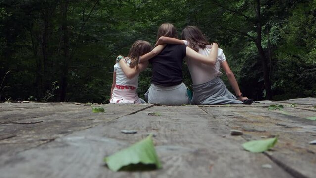 Tres amigas abrazadas y sentadas sobre un puente de madera con un frondoso bosque al fondo