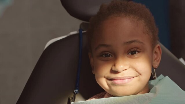 Medium closeup portrait of pretty 6 year old African American girl smiling at camera while sitting in dental chair visiting pediatric dentist