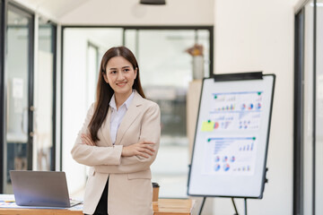 Young asian female accountant standing with arms crossed in front of desk with laptop and financial documents in background in office