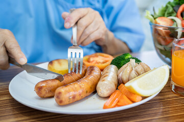 Asian senio woman patient eating breakfast with vegetable healthy food while sitting and hungry on bed in hospital.