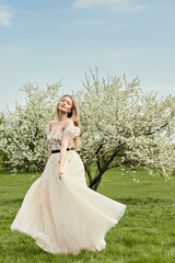 Obraz na płótnie Canvas A young beautiful blonde in a long white dress poses near a cherry blossom in the garden, a spring picturesque landscape