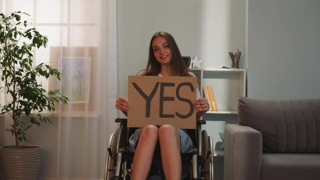 Happy woman sits in wheelchair near houseplant holding poster with word Yes. Young person with disability with paralysis smiles looking in camera