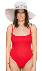 Beautiful brunette woman wearing swimsuit and summer hat skeptic and nervous, frowning upset...