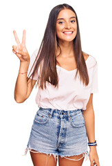Young hispanic woman wearing casual white tshirt smiling looking to the camera showing fingers doing victory sign. number two.