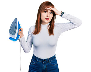 Redhead young woman holding electric steam iron stressed and frustrated with hand on head,...