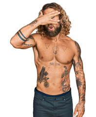 Fototapeta premium Handsome man with beard and long hair standing shirtless showing tattoos peeking in shock covering face and eyes with hand, looking through fingers with embarrassed expression.