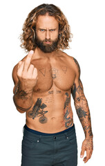 Handsome man with beard and long hair standing shirtless showing tattoos showing middle finger, impolite and rude fuck off expression