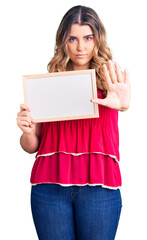 Fototapeta premium Young caucasian woman holding empty white chalkboard with open hand doing stop sign with serious and confident expression, defense gesture