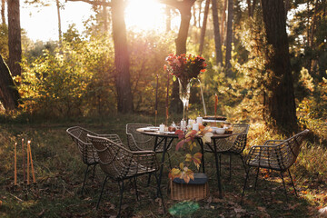 Cozy autumn picnic in the park. Close up of table setting with white plates, cutlery, glass vase with colourful wildflowers, homemade apple pie, maple leaves, burning candles on wooden table outdoors.