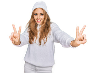 Obraz na płótnie Canvas Young caucasian girl wearing wool sweater and winter cap smiling looking to the camera showing fingers doing victory sign. number two.