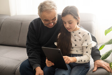 Cute little girl and her handsome grandpa are smiling while sitting on couch at home. Girl is using a tablet.