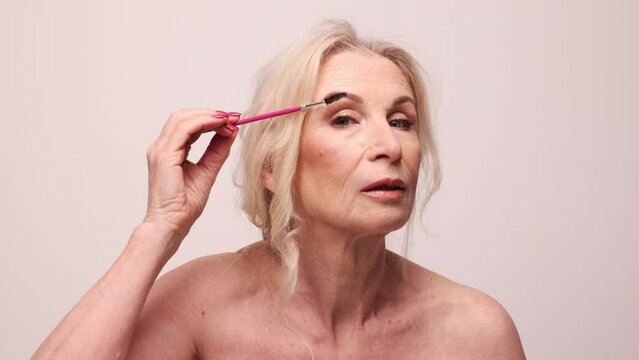 Beautiful middle-aged woman with well-kept healthy skin, nude makeup brushing eyebrows agaisnt grey studio background. Mature old lady. Concept of natural beauty, skin care, cosmetology and cosmetic