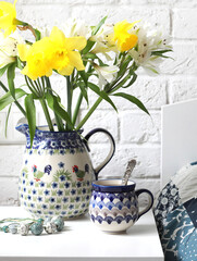 A ceramic mug with tea and a jug with astromelia flowers and daffodils on a white bedside table...