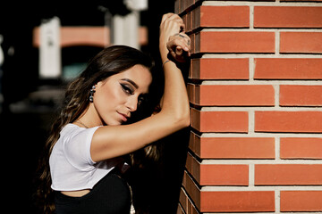 Fototapeta na wymiar A serious woman in profile leaning against a brick wall wearing makeup and jewelry and dressed in casual clothes