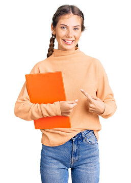 Beautiful caucasian woman with blonde hair holding book smiling happy pointing with hand and finger