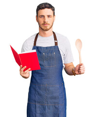 Handsome young man with bear wearing professional baker apron reading cooking recipe book thinking...