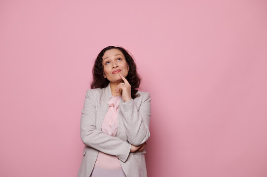 Pensive Middle-aged Woman In Elegant Light Gray Suit, Thoughtfully Looking Up, Holding Finger At Her Chin, Reasoning, Finding Solutions Of Problems, Isolated Pink Color Background With Copy Ad Space