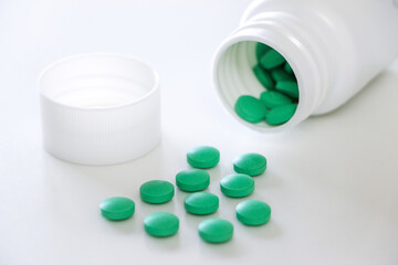 Round green tablets, capsules or pills spilled out of white plastic prescription bottle onto white table with cap. Medicine and pharmaceutical concept.