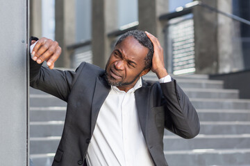 An upset senior African-American man in a suit stands outside an office center, leaning on the wall...