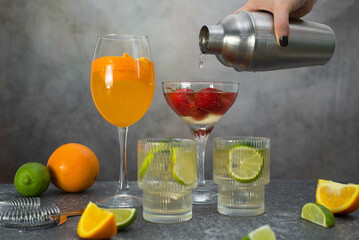 hand woman is holding shaker and pouring coctail in glass. assorted cocktails: two glasses of mojito, orange cocktail, cocktail with strawberries, fruits and shaker on a gray background.