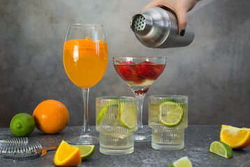 hand woman is holding shaker and pouring coctail in glass. assorted cocktails: two glasses of mojito, orange cocktail, cocktail with strawberries, fruits and shaker on a gray background.