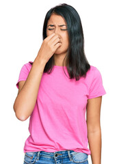 Beautiful asian young woman wearing casual pink t shirt smelling something stinky and disgusting, intolerable smell, holding breath with fingers on nose. bad smell