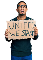 Young african american man holding united we stand banner angry and mad screaming frustrated and furious, shouting with anger looking up.
