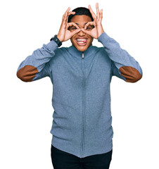 Young handsome hispanic man wearing casual sweatshirt doing ok gesture like binoculars sticking tongue out, eyes looking through fingers. crazy expression.