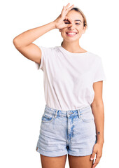 Young beautiful blonde woman wearing casual white tshirt doing ok gesture with hand smiling, eye looking through fingers with happy face.