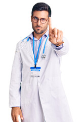 Young hispanic man wearing doctor uniform and stethoscope pointing with finger up and angry expression, showing no gesture