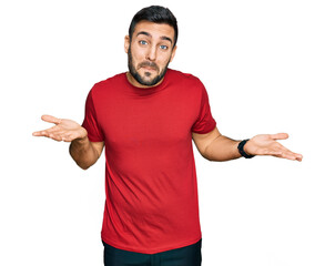 Naklejka premium Young hispanic man wearing casual clothes clueless and confused expression with arms and hands raised. doubt concept.