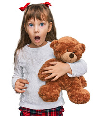 Little caucasian girl kid hugging teddy bear stuffed animal scared and amazed with open mouth for...