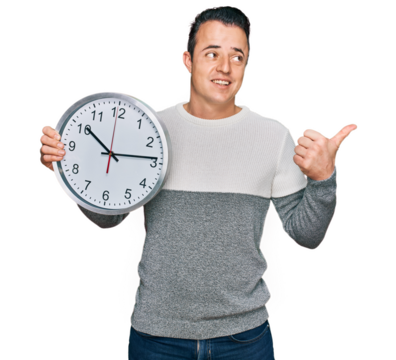 Handsome young man holding big clock pointing thumb up to the side smiling happy with open mouth
