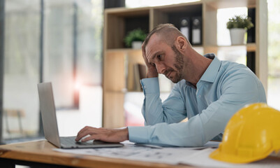 Fototapeta premium Bored and tired caucasian male engineer or architect sits at his desk, hands on his chin, looking at the camera, feeling no energy to work, overworked, and lacking passion.
