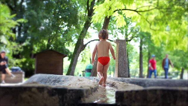 Back of cute child wearing red bathing suit brief outside at park during sunny summer day. Lifestyle activity of small boy enjoying nature barefoot
