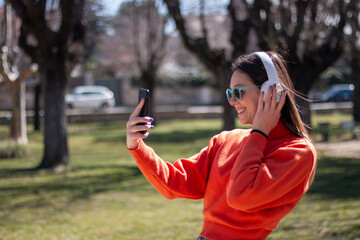 Young brunette woman smiling with headphones, sunglasses and mobile phone in her hand, dressed in orange sweater and jeans