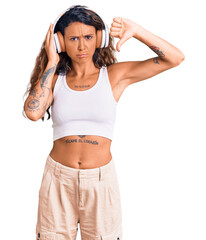 Young hispanic woman with tattoo listening to music using headphones with angry face, negative sign showing dislike with thumbs down, rejection concept