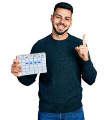 Young hispanic man with beard holding rainy weather calendar smiling with an idea or question pointing finger with happy face, number one