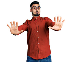 Young hispanic man with beard wearing business shirt and glasses afraid and terrified with fear expression stop gesture with hands, shouting in shock. panic concept.