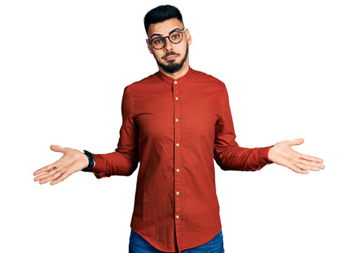 Young hispanic man with beard wearing business shirt and glasses clueless and confused expression with arms and hands raised. doubt concept.