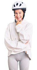 Beautiful brunette young woman wearing bike helmet and sporty clothes looking confident at the camera smiling with crossed arms and hand raised on chin. thinking positive.