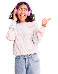 Young beautiful mixed race woman listening to music using headphones pointing thumb up to the side...