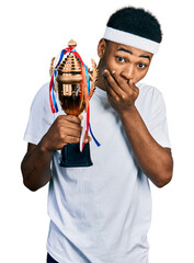 Young african american man holding winner trophy covering mouth with hand, shocked and afraid for...